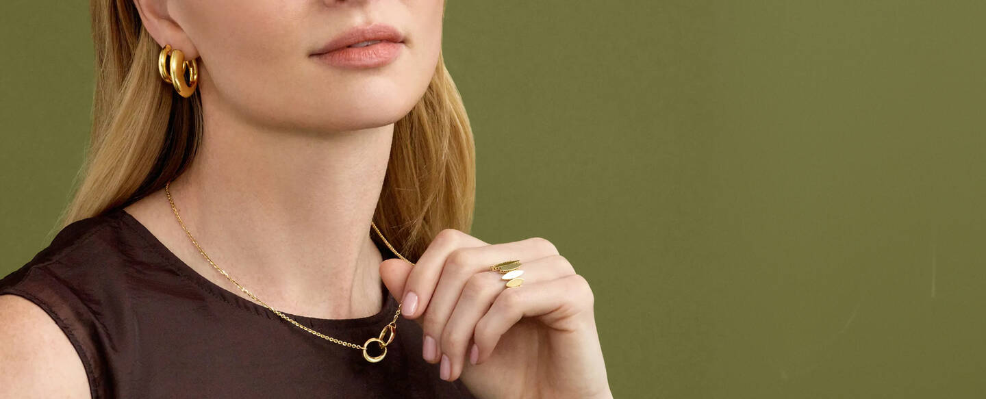 A woman wearing a brown tank top adjusts a yellow gold pendant necklace while wearing bold gold hoop earrings and a yellow gold ring. 