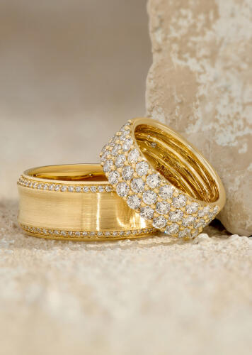 A yellow gold band in a satin finish features a perimeter of round diamonds. A diamond anniversary ring leans against the wide band.