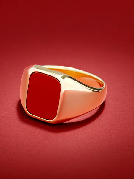 A yellow gold signet ring with a square red center is shown on a textured red surface.
