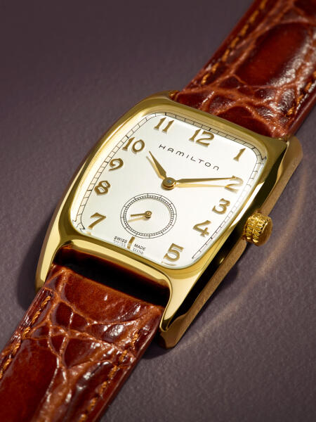 A Hamilton dress watch with gold case and brown leather strap rests on a textured purple surface.