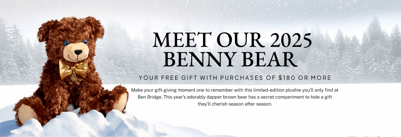 A limited edition 2025 Benny Bear plushie sits on a little mound of show with a snowy winter landscape in the background. 