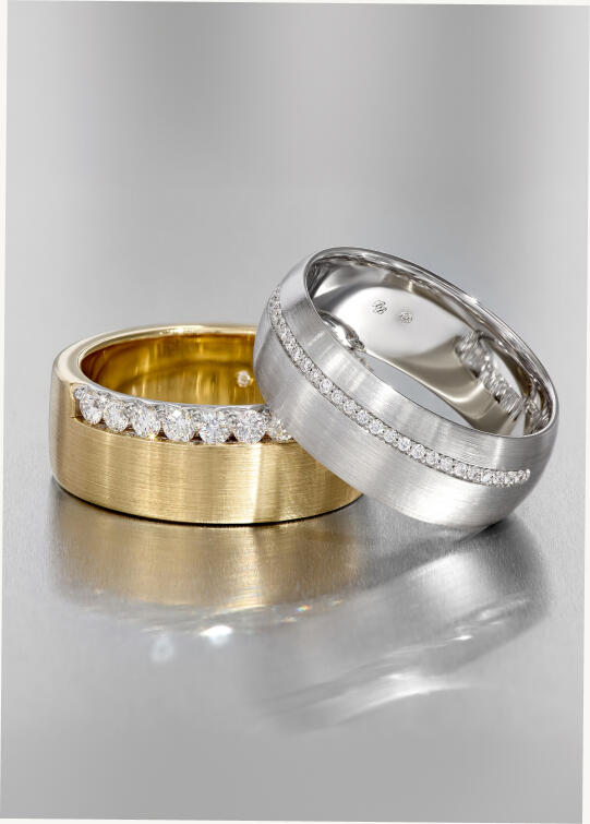 Two diamond wedding bands with round diamonds, one in yellow gold and the other in white gold on a smooth reflective surface.