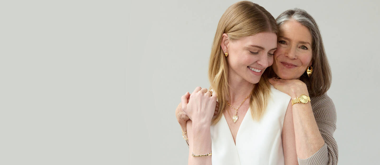 A mother embraces her daughter, both smiling and wearing yellow gold jewelry.