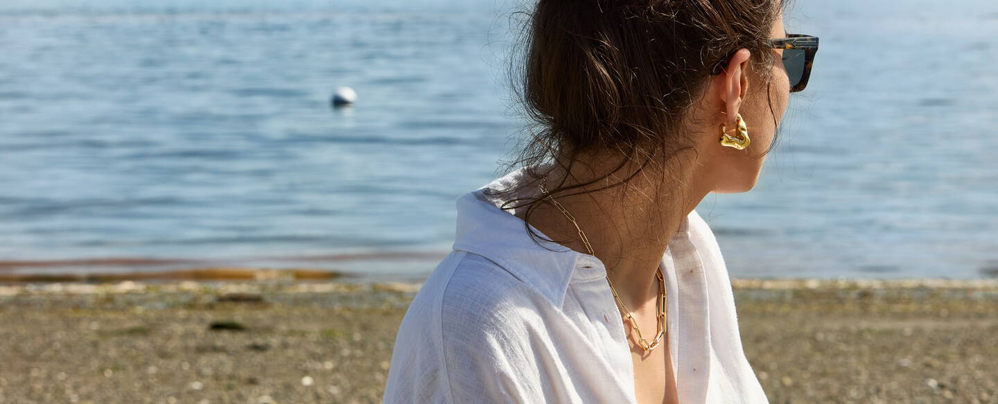 A woman wearing sunglasses and gold jewelry looks happily out over the ocean waters.