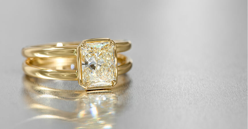A solitaire diamond engagement ring set in in yellow gold is reflected off the surface of a metal table.