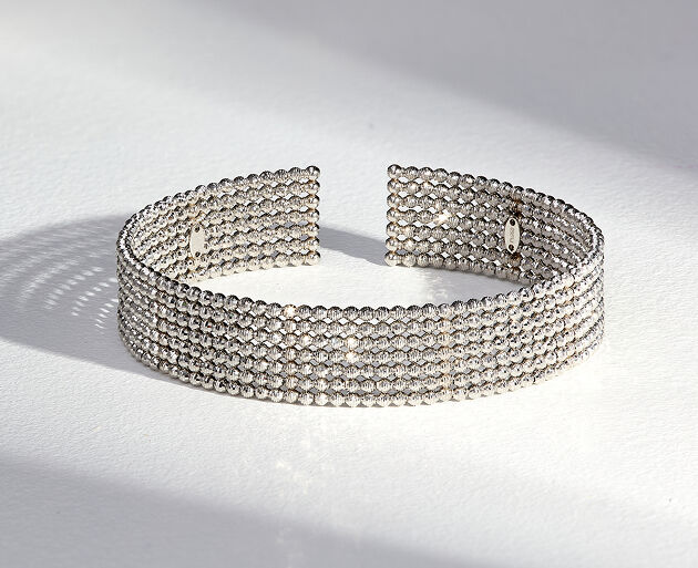 A platinum bracelet with several rows of diamond cut beads casting a shadow across a smooth light gray surface.