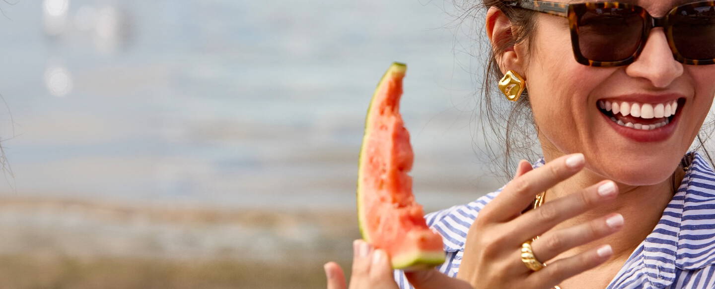 Woman wearing sunglasses is eating watermelon and laughing at the beach while wearing yellow gold jewelry.
