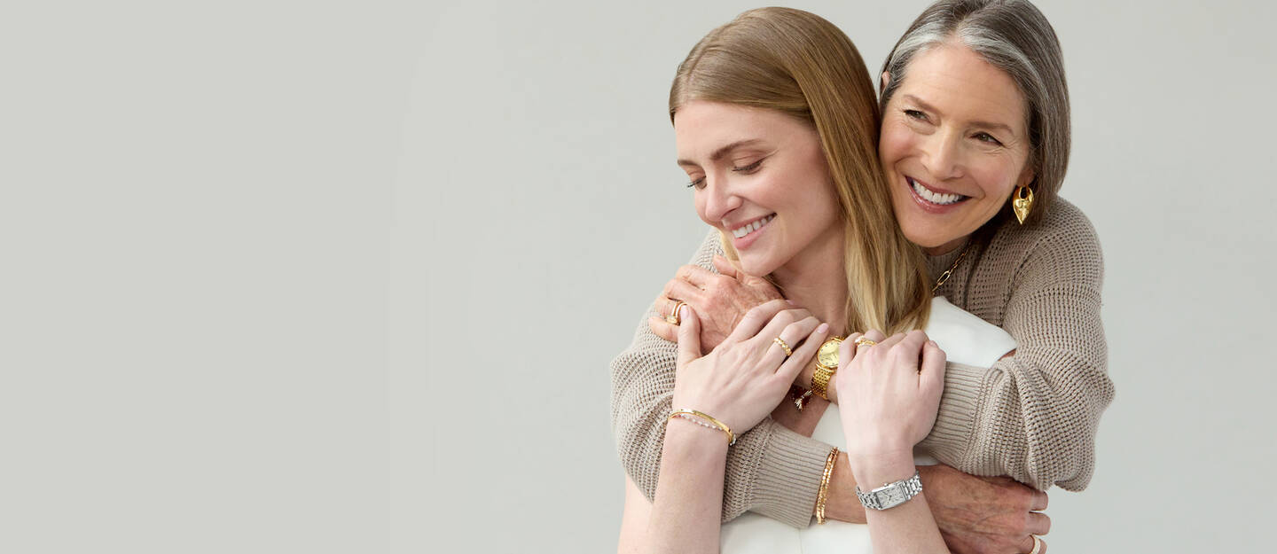 A mother embraces her daughter, both smiling and wearing yellow gold jewelry. 