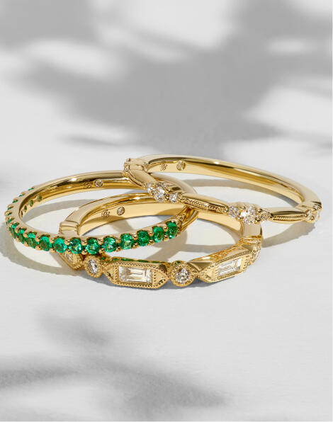 Three yellow gold bands featuring diamonds of varying shapes and natural green gemstones. 