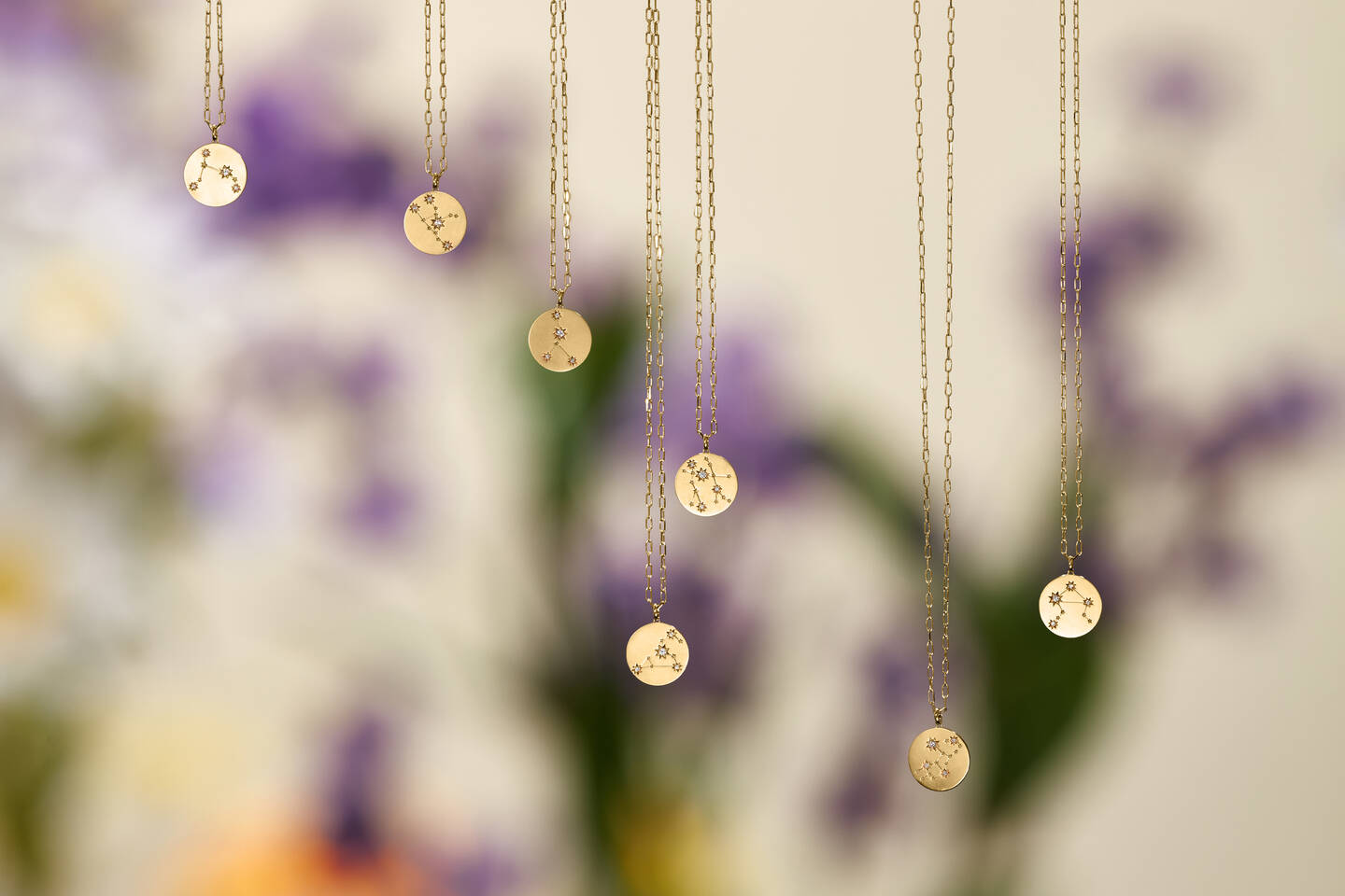 Zodiac pendants on gold chains stand out against a blurred purple, green and white background. 