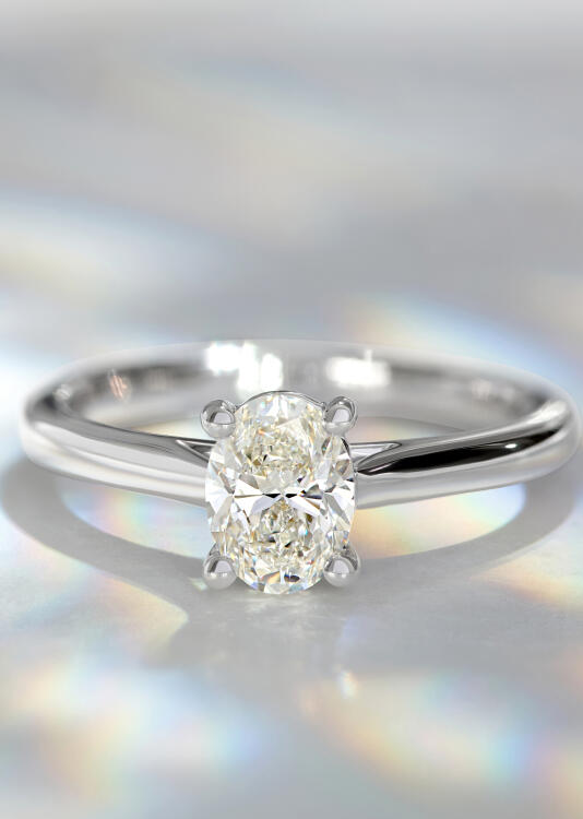 A solitaire diamond engagement ring in white gold rests on its side on a smooth grayish-white surface.