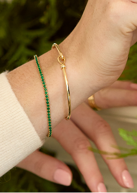 Closeup of a woman’s wrist highlights two bracelets, one of yellow gold and the other featuring green gemstones.