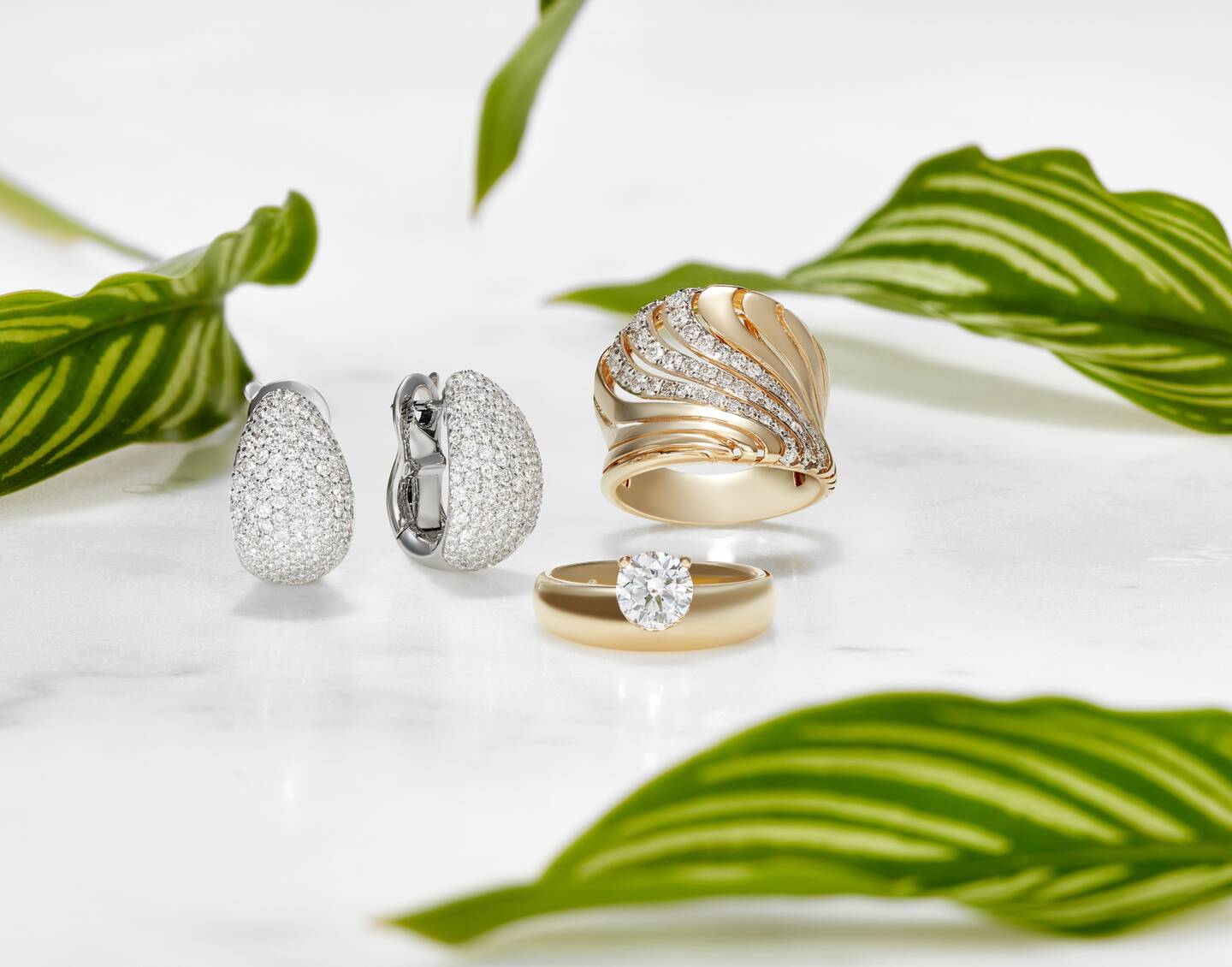 earrings made with white gold and diamonds next to a solitaire diamond ring in yellow gold and a liquid shaped gold and diamond ring. 