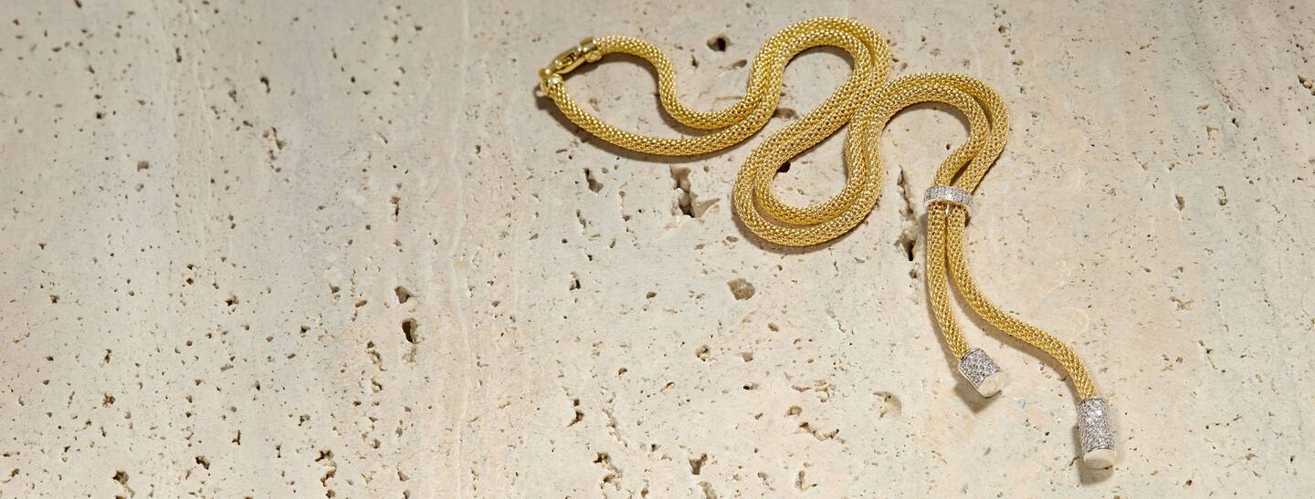 A bolo-style yellow gold necklace features white gold and diamond accents and rests on a porous stone surface.