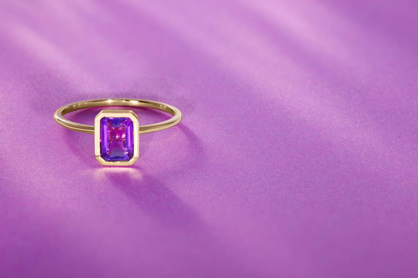 Emerald cut purple amethyst gemstone ring in 14k yellow gold shown against a purple background. 