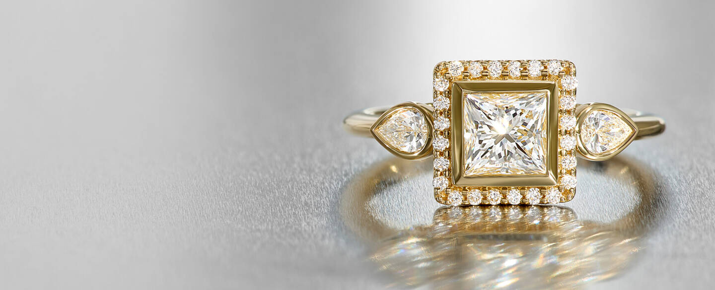 A yellow gold diamond engagement ring with a halo setting and two pear-shaped diamonds in bezel settings reflects on a silver surface.