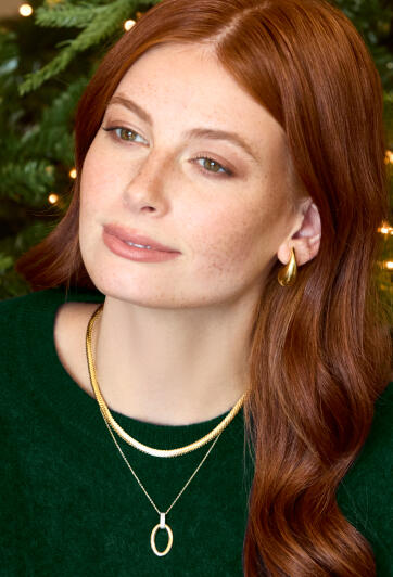 A smiling woman is dressed in a green sweater with gold earrings and a gold necklace.