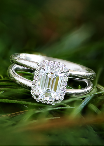 A double shank diamond halo engagement ring in white gold rests on a bed of evergreen leaves. 