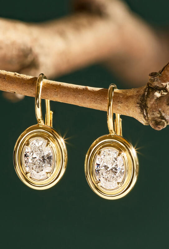 Bezel set oval diamond earrings in yellow gold dangle from a Christmas tree branch.