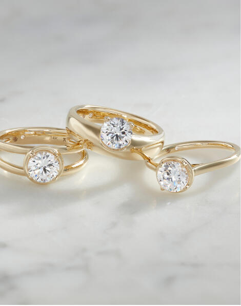Three solitaire diamond engagement rings in yellow gold on a white marble surface.