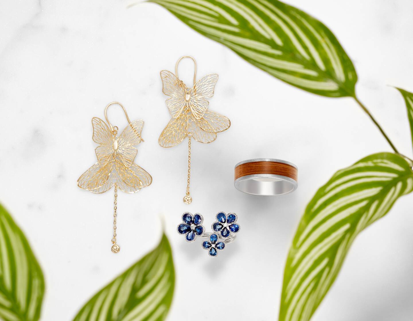 Butterfly shaped earrings, flower shaped sapphire ring and a silver ring with a wooden center against a white background. 