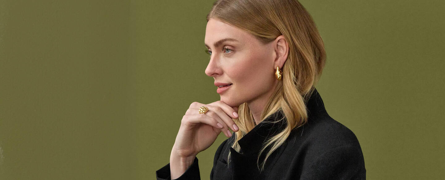 A woman wearing yellow gold hoops gazes thoughtfully toward the right with her hand on her chin.