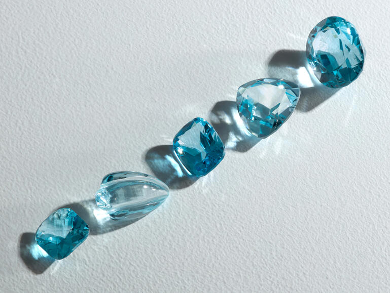 Aquamarine gemstones in varying shapes and shades of blue reflect across a textured white surface.