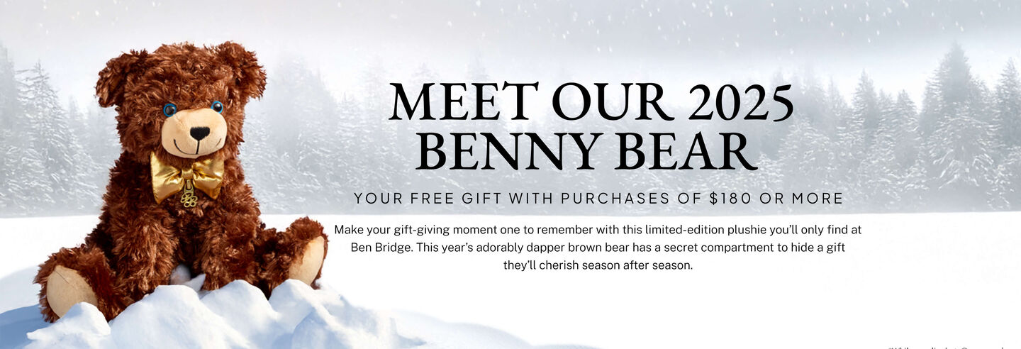 Benny bear plushie with a gold bowtie sits atop a mound of snow amid a wintery scene.