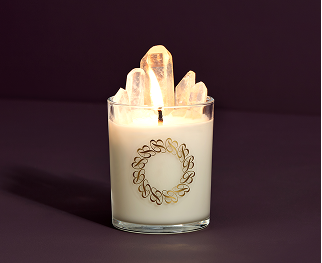 A cream-colored candle with a gold Ben Bridge logo and charged with Reiki crystals glows against a purple background.