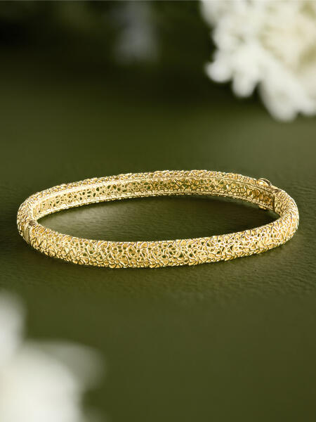 A yellow gold bangle rests on a green surface surrounded by white flowers.