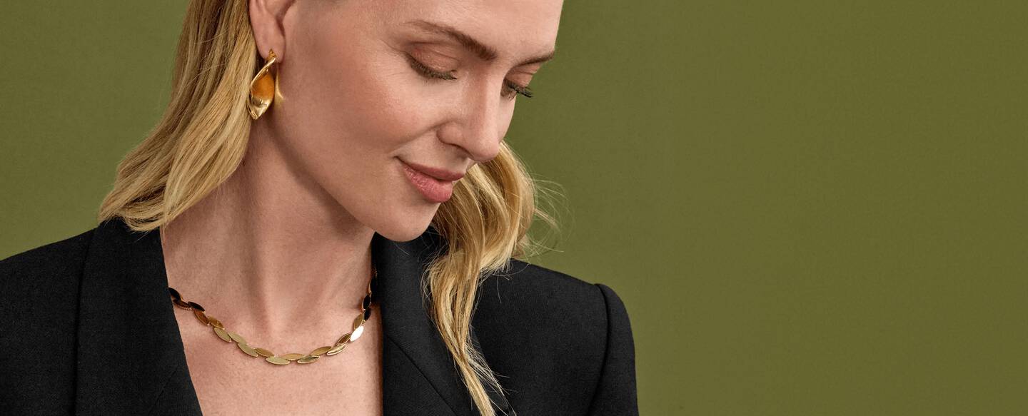 A woman wearing a black blazer and yellow gold necklace with matching earrings.