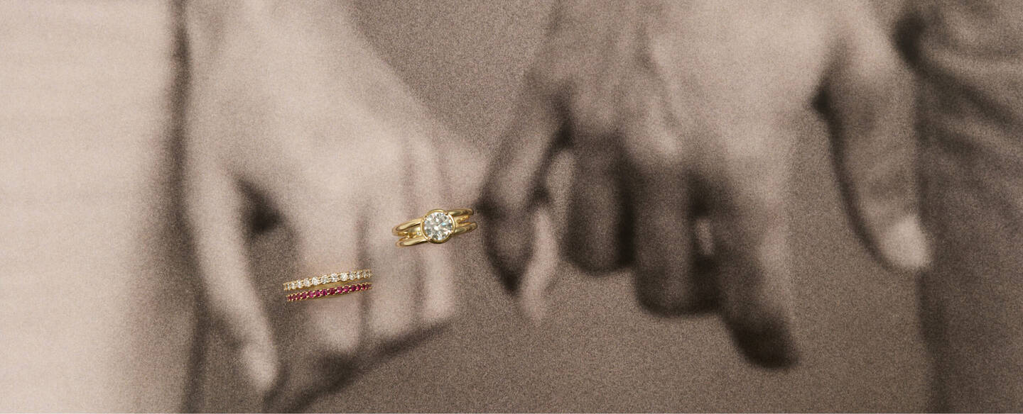 Two hands interlock in a soft focus that gives a watercolor-like feel to the scene. One hand is wearing an engagement ring and gemstone anniversary bands.