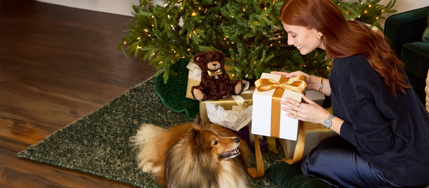 A woman is holding a gift near a tree decorated for the holiday while a dog watches.