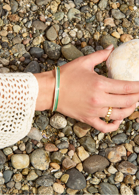 A woman&rsquo;s hand reaches for a stone. She is wearing a yellow gold bracelet