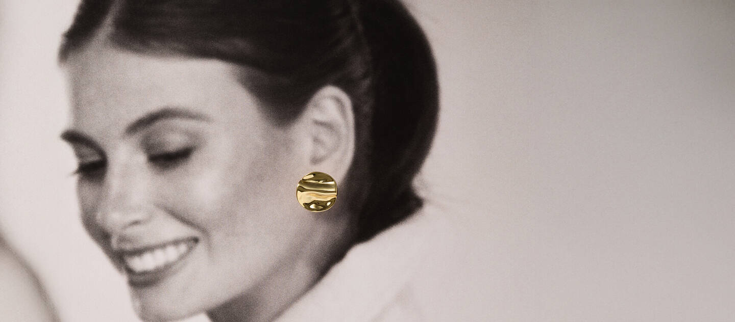 Black and white scene of a woman smiling. The only color is a yellow gold pair of disc-shaped earrings.