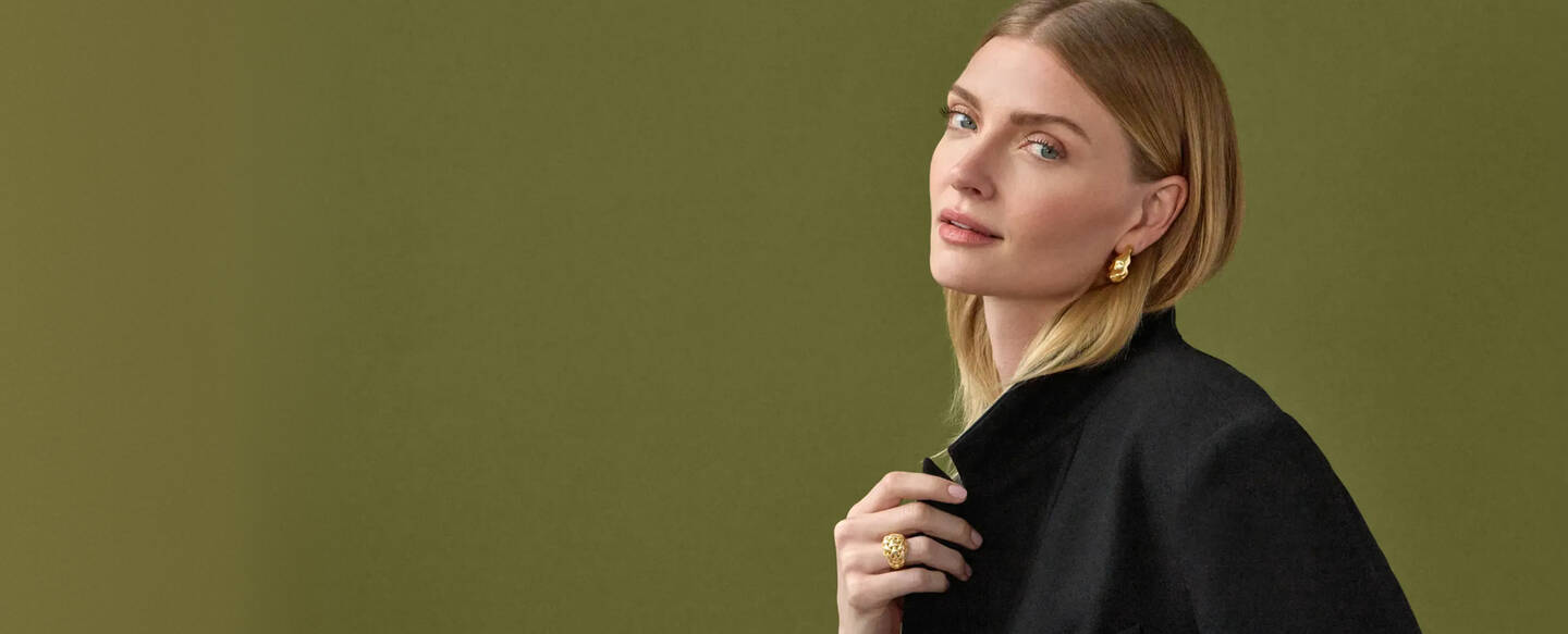 A woman in yellow gold earrings smiles slightly while adjusting the collar of her black coat.