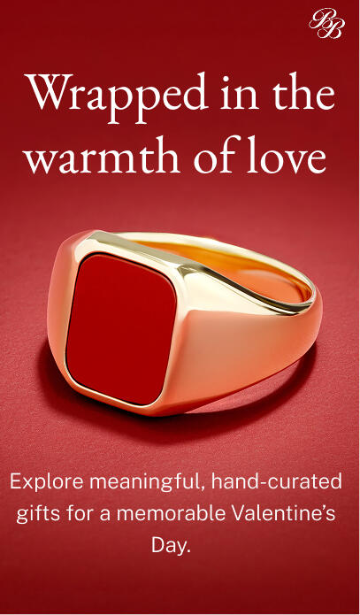 A yellow gold signet ring with a square red center is shown on a textured red surface.