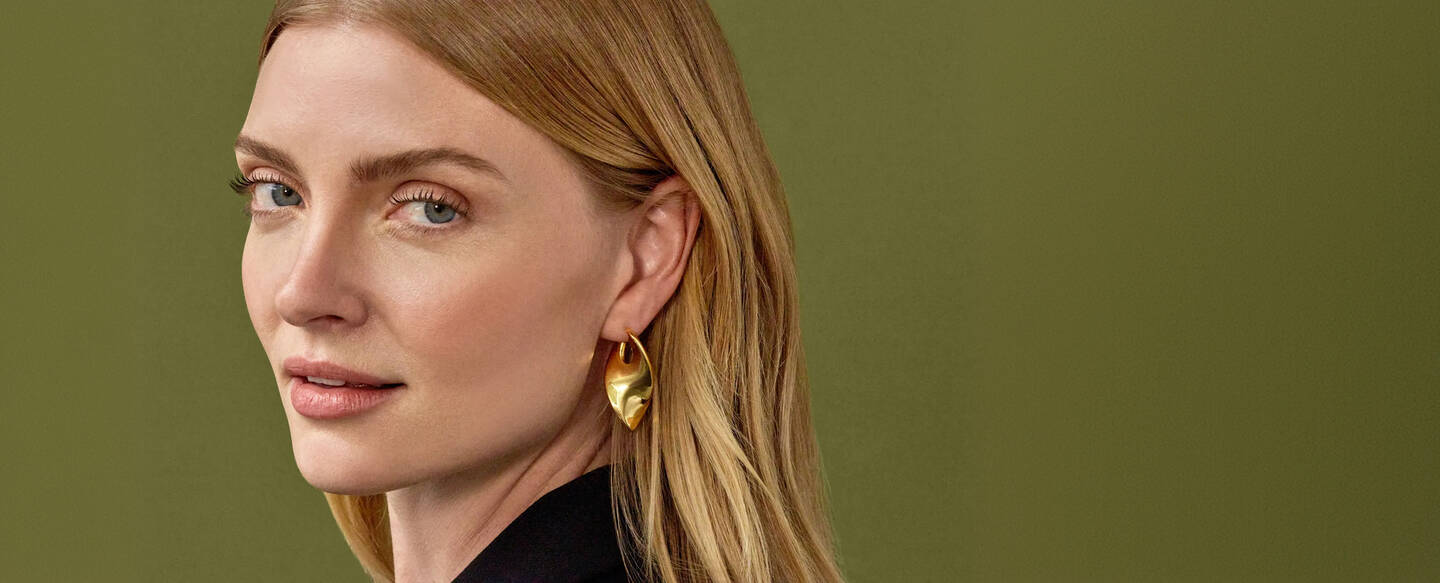 A woman with long hair glances to her right. She is wearing bold yellow gold earrings.