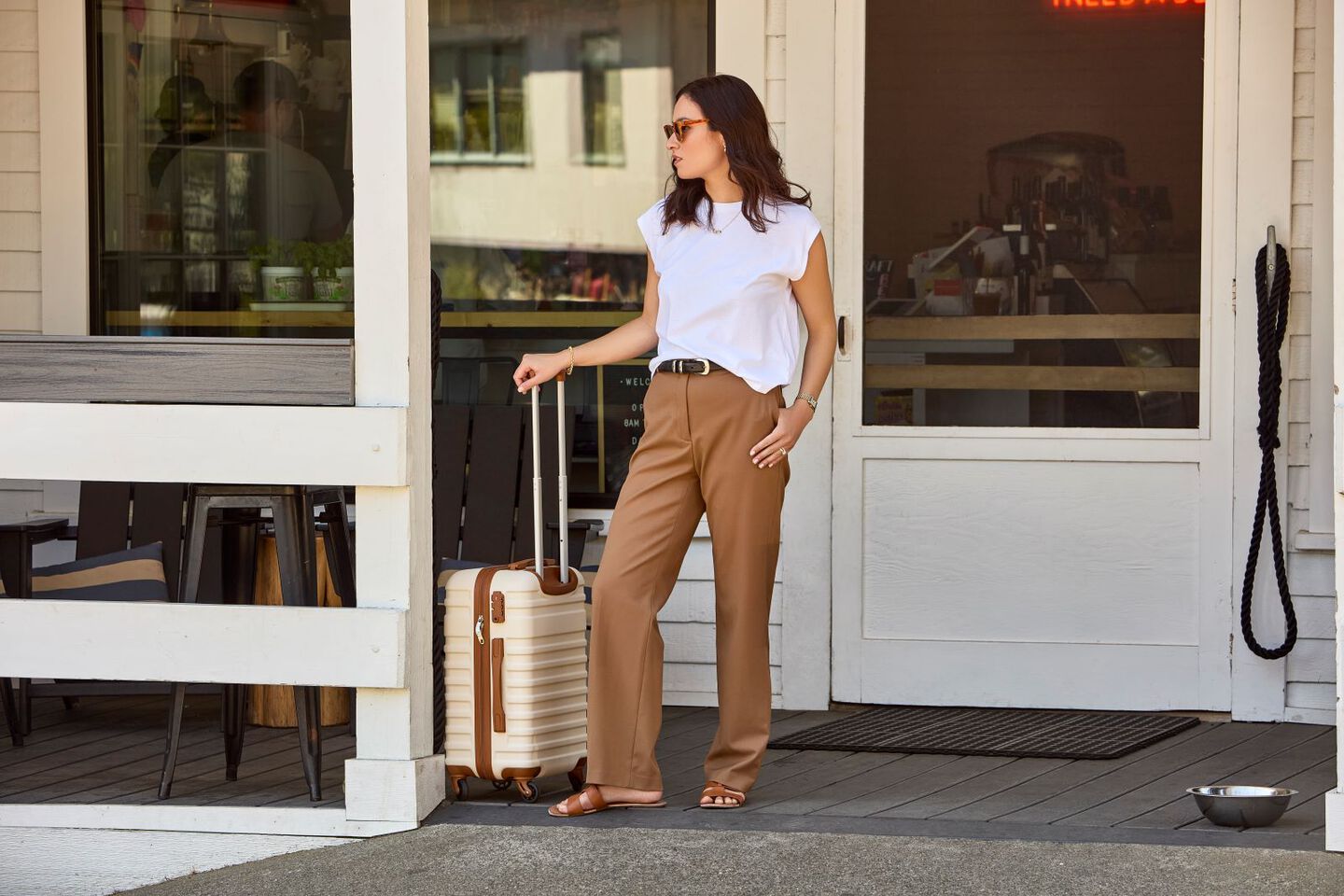 a woman stands outside a white home with a rolling suitcase in her hand. 