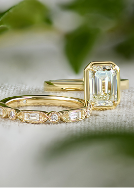 A solitaire engagement ring in yellow gold behind a diamond band set in yellow gold rests on a textured surface.