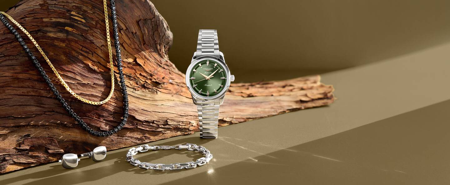 A yellow gold and black necklace are draped over a driftwood log while a stainless steel sports watch with a green dial stands upright next to a silver bracelet.