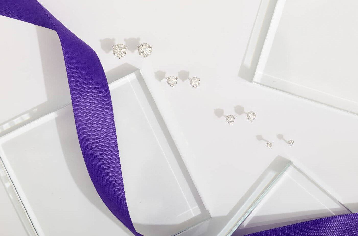 Diamond stud earring in various sizes against a while background with a flowing purple ribbon 