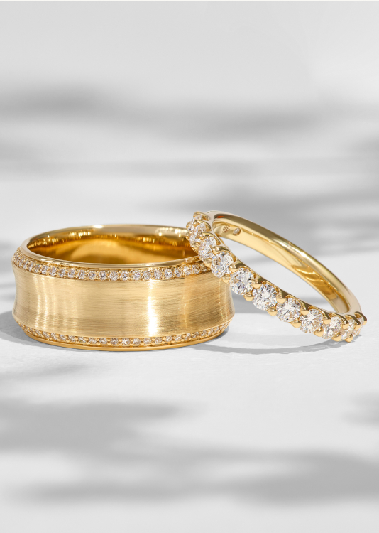 A yellow gold band in a satin finish features a perimeter of round diamonds. A diamond anniversary ring leans against the wide band.