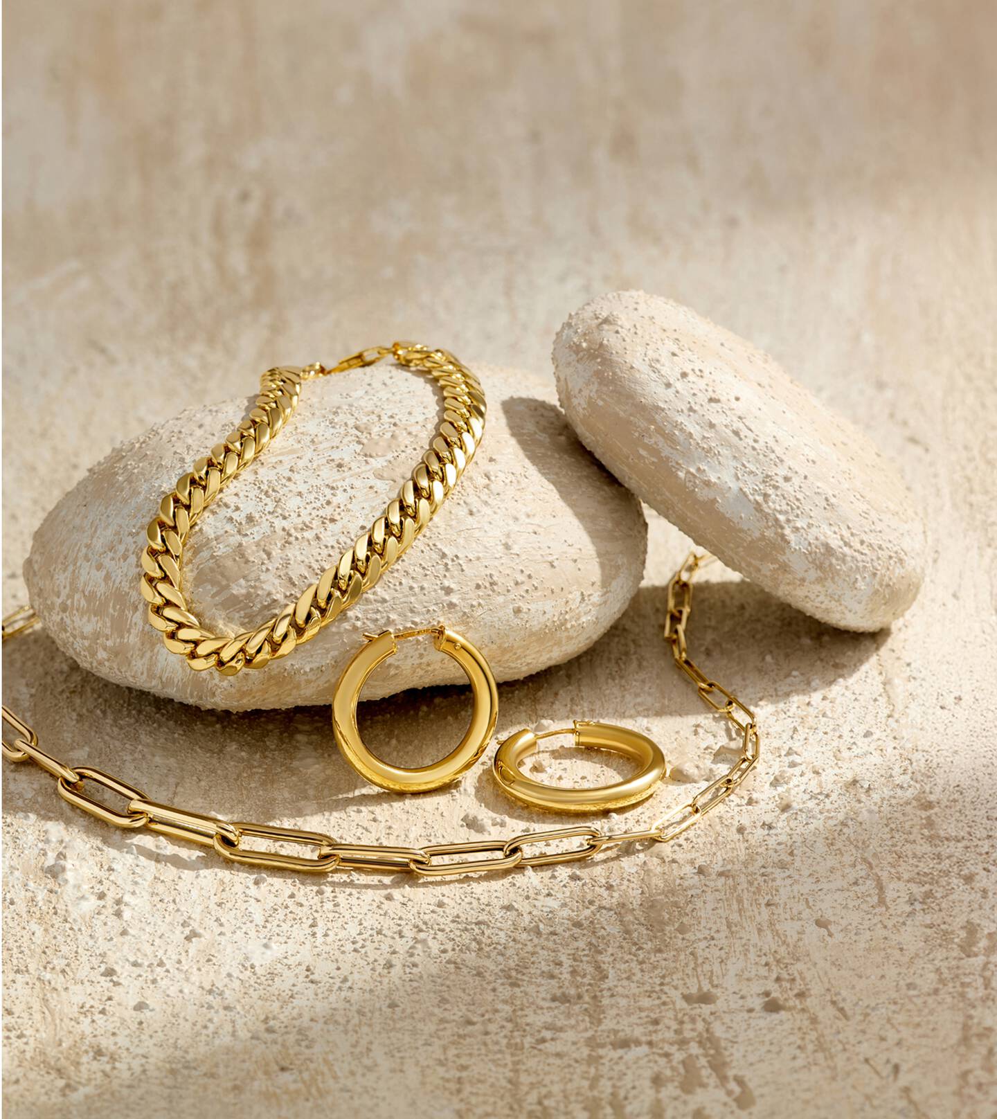 14k yellow gold bracelet, hoop earrings and necklace rest on white rocks that cast shadows over the surface of a white stone table. 