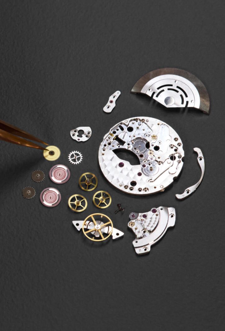 Parts of a dismantled mechanical watch movement spread out on a gray surface. 
