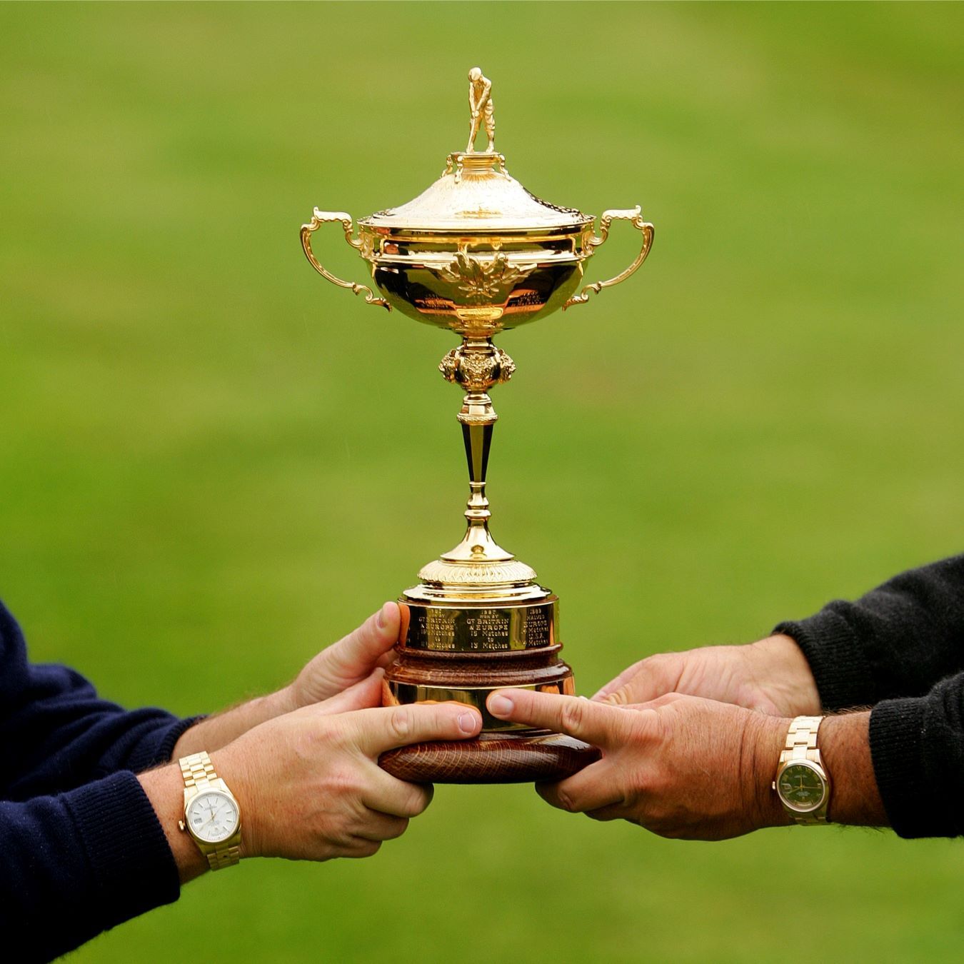 Two hands wearing Rolex watches hold the Ryder Cup trophy. 