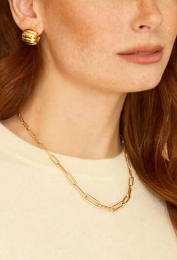 A woman in gold earrings and a cream colored sweater wearing a yellow gold link necklace.