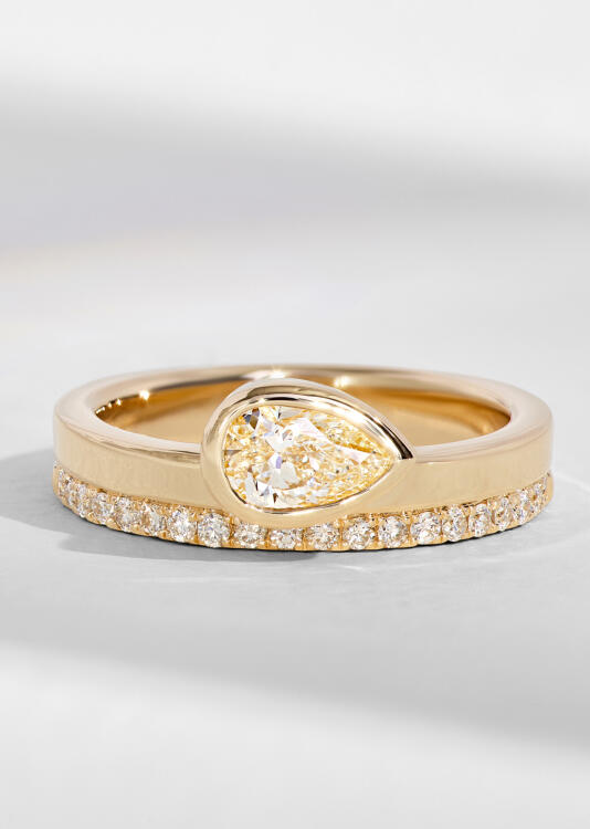 A yellow gold engagement ring features a pear shaped diamond in a bezel setting alongside a diamond wedding band.