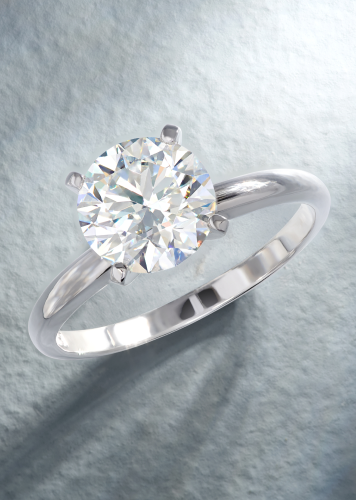 A solitaire diamond engagement ring in white gold balances on a smooth shank on a gray textured surface.