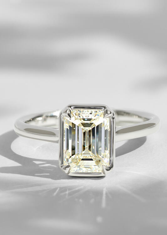 An emerald cut solitaire diamond engagement ring in while gold with a bezel setting.